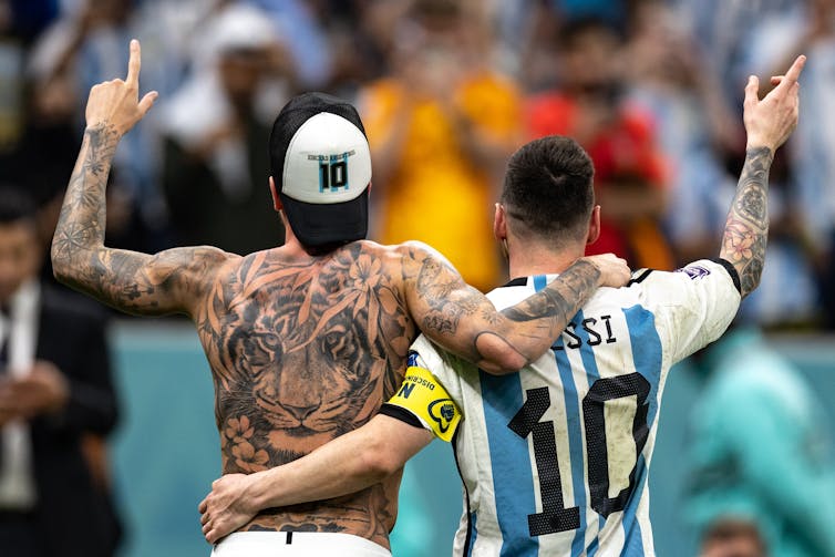 A large tiger tattoo is visible on the bare back of one soccer player, while another player's arm tattoo can be seen as they raise their right hands after a victory.