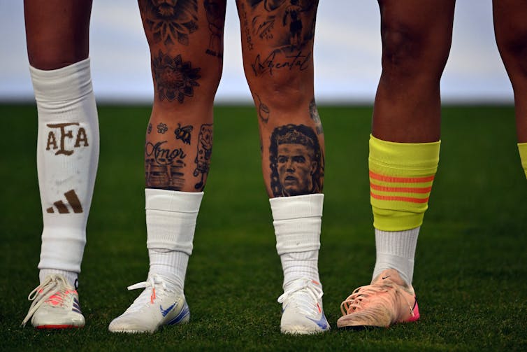 Four players' legs on a soccer field, two of them covered in tattoos.