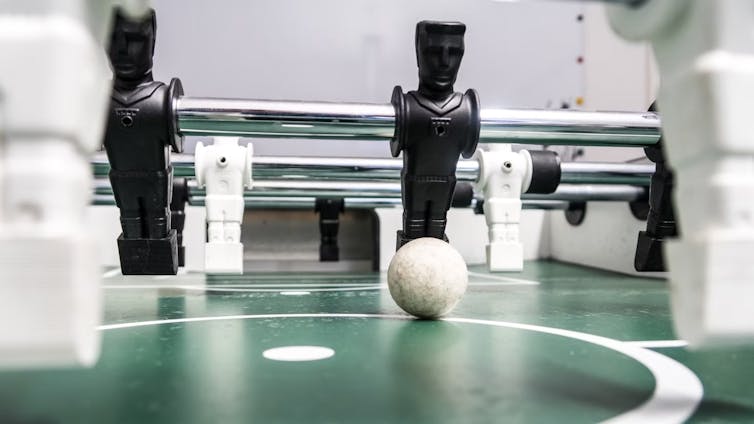 A foosball set with toy figures on metal rods used to propel a small football, a row of black figures on strike, white figures foregrounded.